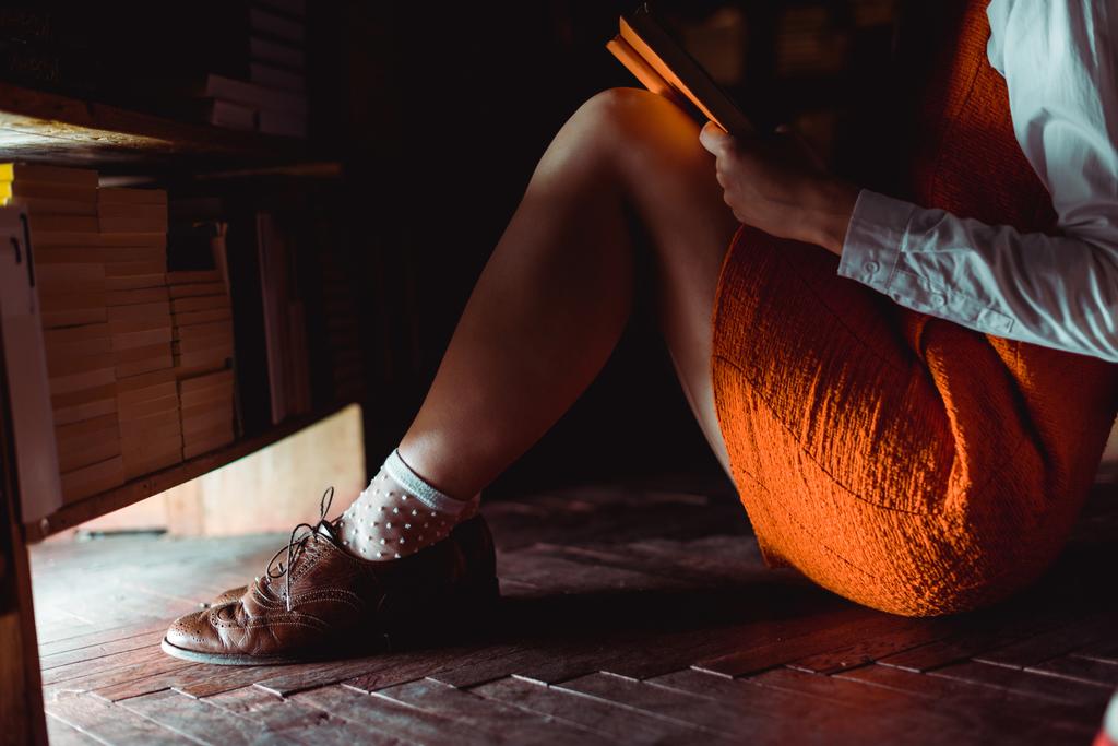 περικομμένη θέα της γυναίκας που κάθεται στο ξύλινο πάτωμα και κρατώντας το βιβλίο στη βιβλιοθήκη  - Φωτογραφία, εικόνα
