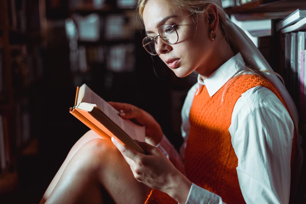 όμορφη και ξανθιά γυναίκα σε γυαλιά διαβάζοντας βιβλίο στη βιβλιοθήκη  - Φωτογραφία, εικόνα