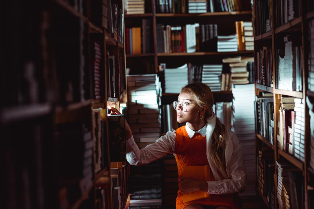 mooie en blonde vrouw in glazen en oranje jurk op zoek weg in de bibliotheek  - Foto, afbeelding