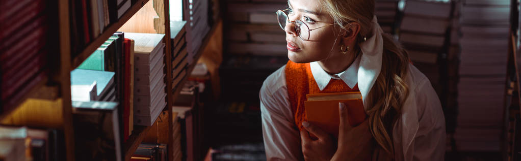 panoramic shot of beautiful woman in glasses holding book and looking away in library  - Photo, Image