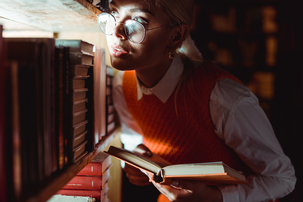 bonita mujer en gafas sosteniendo libro y mirando hacia otro lado en la biblioteca
  - Foto, Imagen