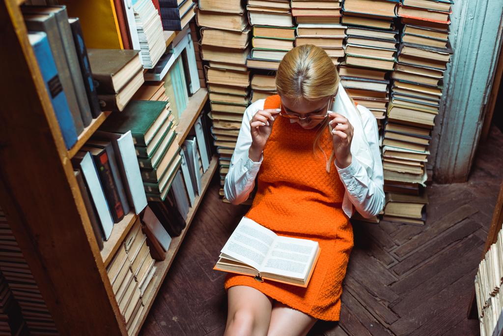 όμορφη γυναίκα σε πορτοκαλί φόρεμα κάθεται στο πάτωμα και διαβάζοντας το βιβλίο στη βιβλιοθήκη - Φωτογραφία, εικόνα