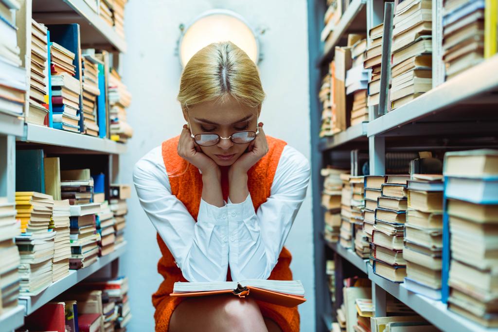 piękna i blond kobieta w pomarańczowej sukni i okulary czytanie książki w bibliotece  - Zdjęcie, obraz