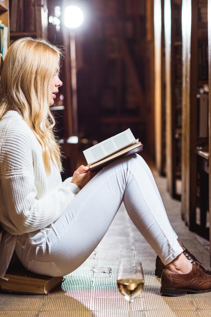 Zijaanzicht van blonde vrouw lezen boek en zittend op de vloer in de bibliotheek  - Foto, afbeelding
