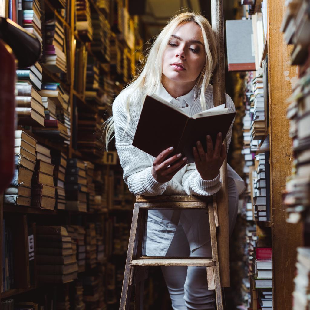 blond i ładna kobieta w białym sweter czytanie książki w bibliotece  - Zdjęcie, obraz