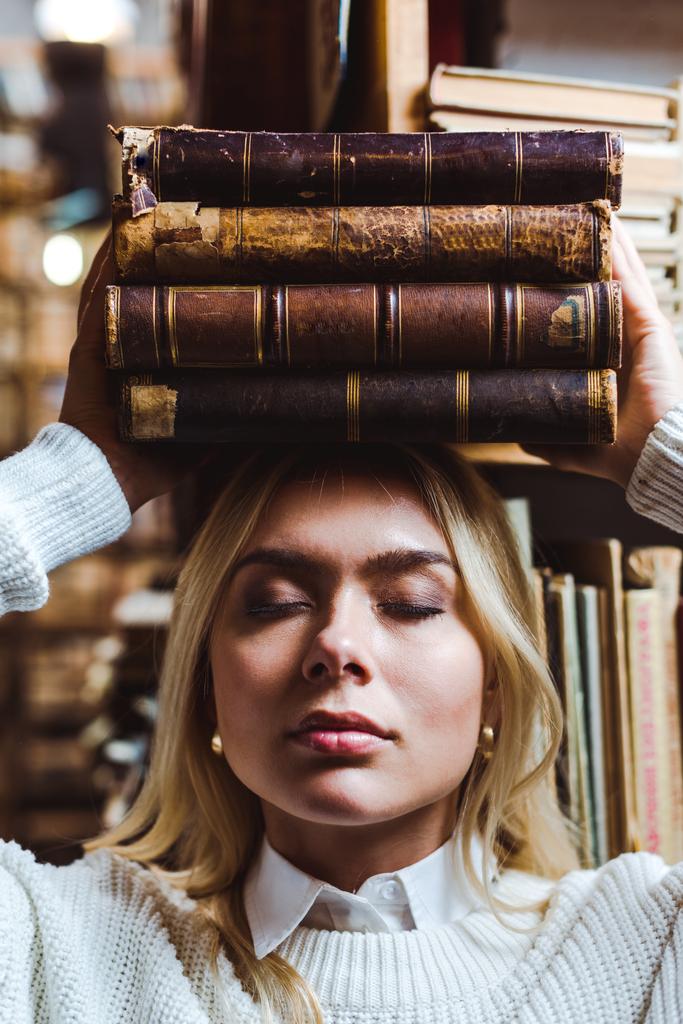 ξανθιά και όμορφη γυναίκα με κλειστά μάτια κρατώντας βιβλία στη βιβλιοθήκη  - Φωτογραφία, εικόνα