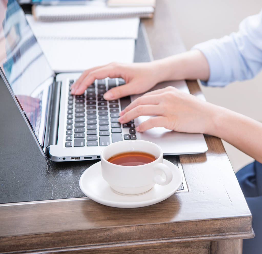 Concepto de negocio. Mujer de camisa azul escribiendo en la computadora con café en la mesa de la oficina, retroiluminación, efecto de deslumbramiento solar, primer plano, vista lateral, espacio para copiar
 - Foto, imagen