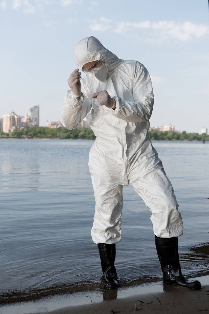 πλήρους μήκους θέα του επιθεωρητή νερού σε προστατευτικά κοστούμια και λατέξ γάντια λαμβάνοντας δείγμα νερού - Φωτογραφία, εικόνα
