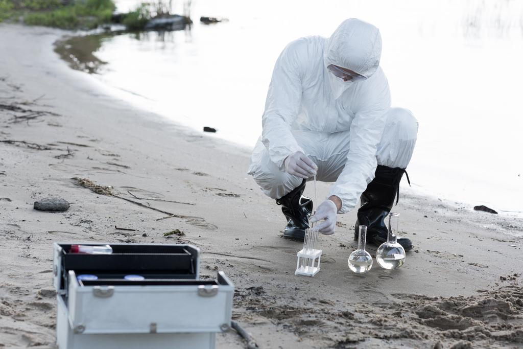 Wasserinspektor in Schutzanzügen und Atemschutzmaske entnimmt Wasserproben am Fluss - Foto, Bild
