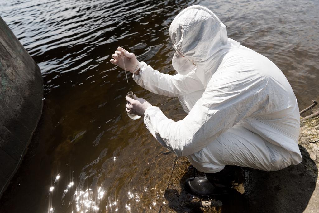 water inspecteur in beschermende kostuum, latex handschoenen en gasmasker nemen watermonster op rivier - Foto, afbeelding
