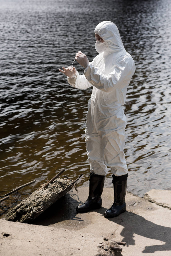 volledige lengte weergave van water inspecteur in beschermende kostuum, latex handschoenen en gasmasker nemen watermonster op rivier - Foto, afbeelding