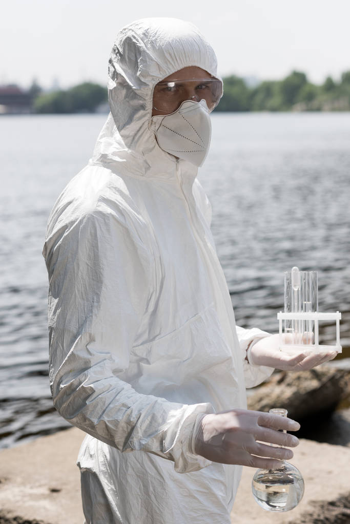 water inspector in protective costume, latex gloves and respirator holding flask and test tubes with water samples at river - Photo, Image