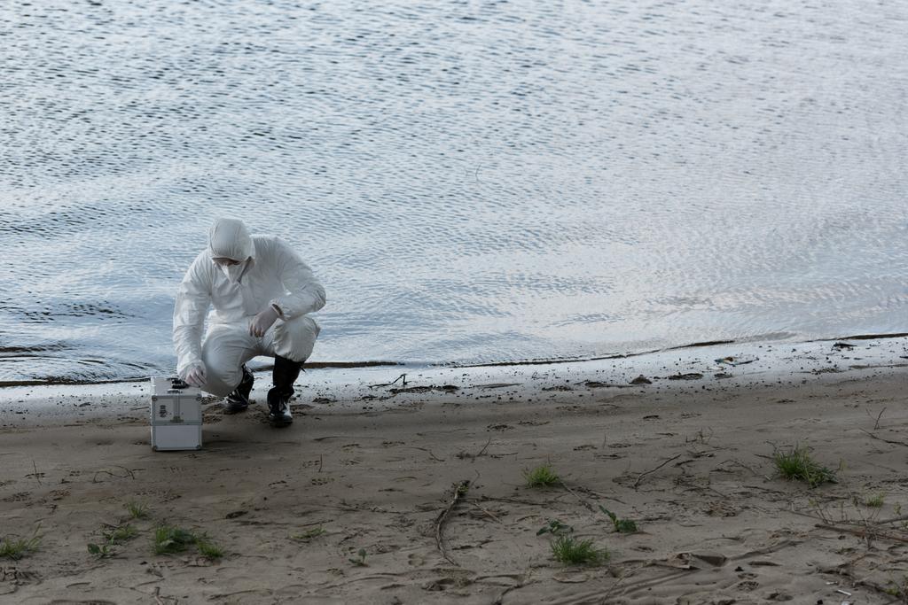 инспектор по воде в защитном костюме и респираторе с инспекционным комплектом, сидящим на берегу реки
 - Фото, изображение