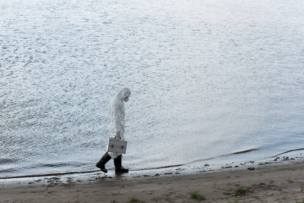 Ansicht des Wasserinspektors in Schutzkleidung und Atemschutzmaske mit Inspektionskoffer beim Spazierengehen an der Flussküste - Foto, Bild