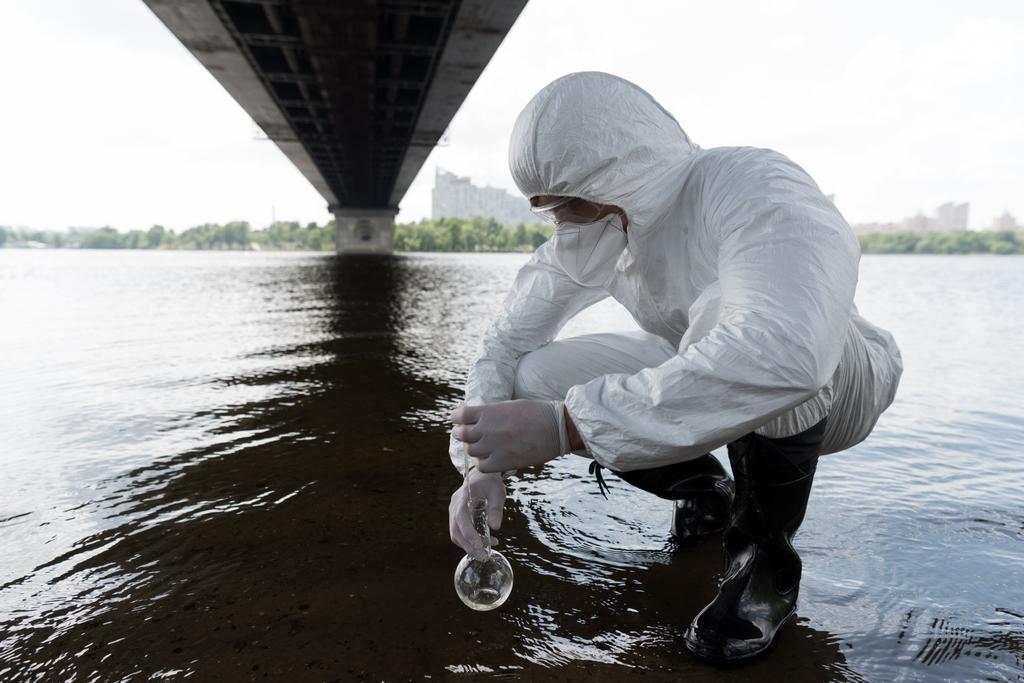 フラスコを保持し、川から水のサンプルを取る保護衣装の水検査官 - 写真・画像