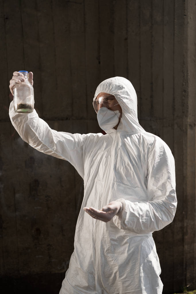 еколог у захисному костюмі та респіраторі, що тримає пляшку з ґрунтовим зразком
 - Фото, зображення