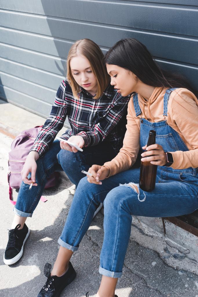 pretty friends smoking cigarettes, holding beer, sitting and looking at smartphone   - Photo, Image