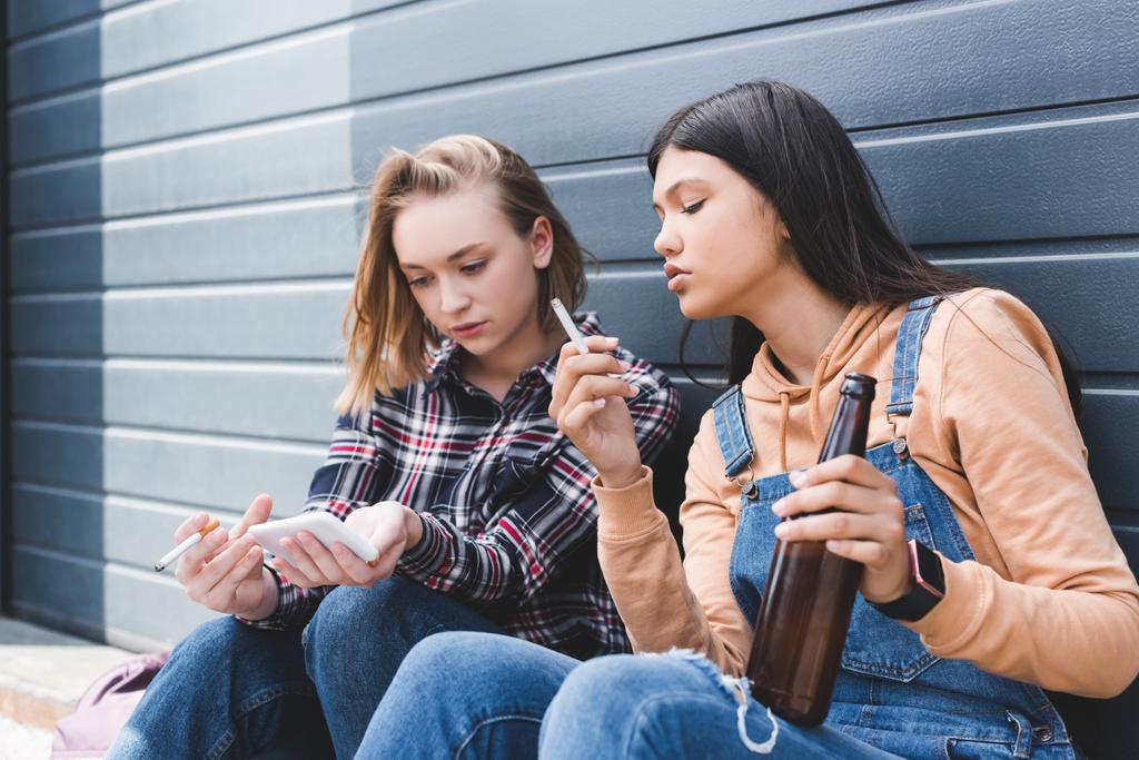 καλοί φίλοι καπνίζουν τσιγάρα, κρατώντας μπύρα, καθιστός και κοιτάζοντας smartphone   - Φωτογραφία, εικόνα