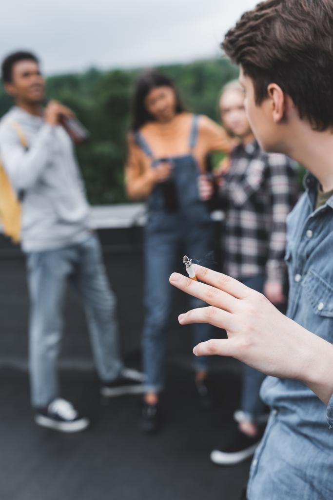 selective focus of teenager smoking cigarette with friends on roof  - Photo, Image