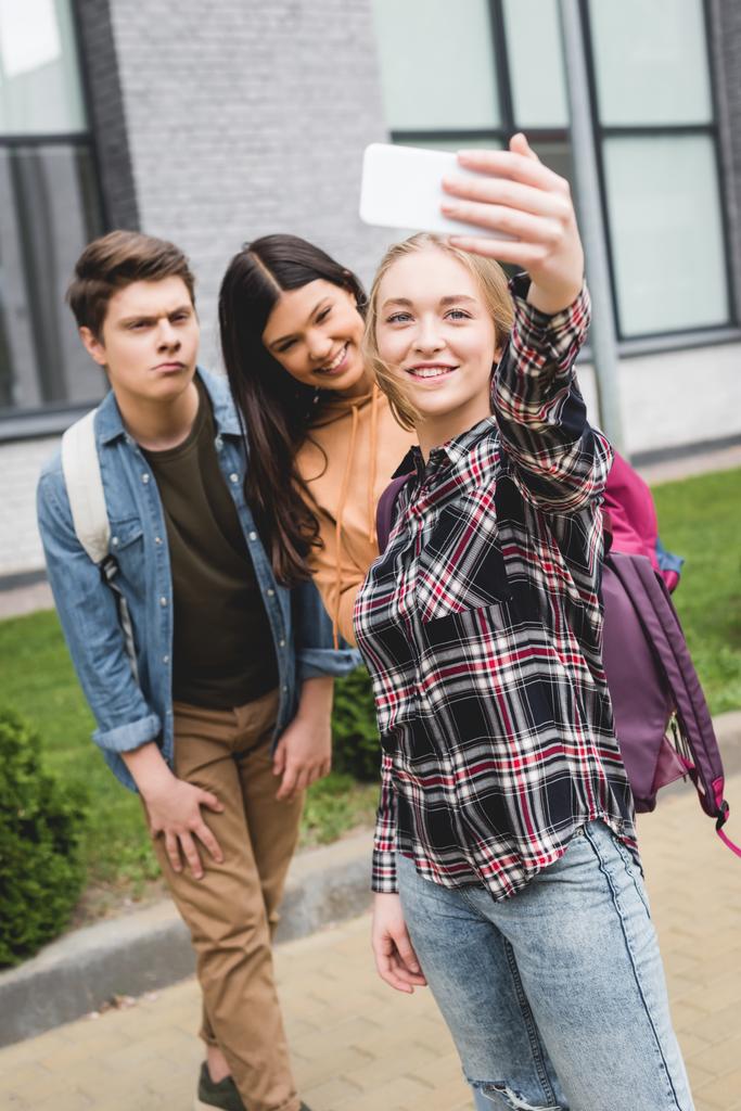 Jugendliche halten Smartphone in der Hand, machen Selfie und lächeln draußen  - Foto, Bild