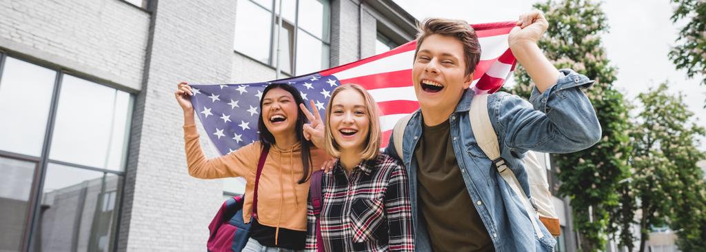 πανοραμική φωτογραφία χαρούμενους εφήβους που χαμογελούν, κρατώντας αμερικάνικη σημαία και κοιτάζοντας την κάμερα  - Φωτογραφία, εικόνα