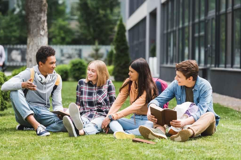 uśmiechnięci nastolatkowie siedzący na trawie, rozmawiając i trzymając książki  - Zdjęcie, obraz