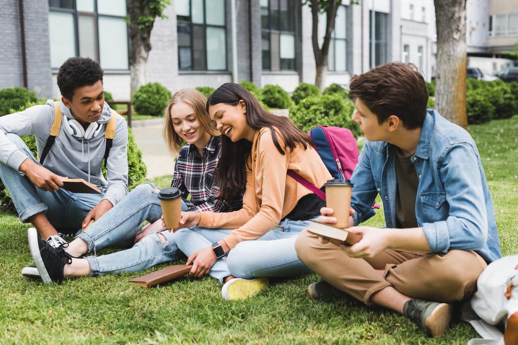 Lächelnde Teenager, die auf Gras sitzen, reden, Bücher und Pappbecher in der Hand halten  - Foto, Bild