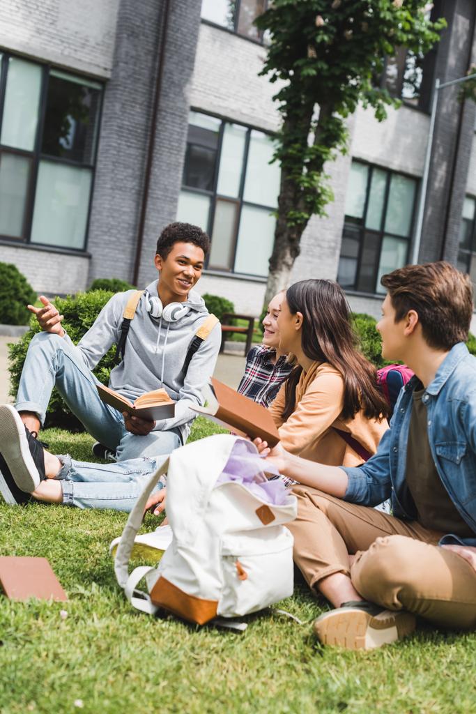 uśmiechnięci i szczęśliwi nastolatkowie siedzący na trawie, rozmawiając, trzymając książki  - Zdjęcie, obraz