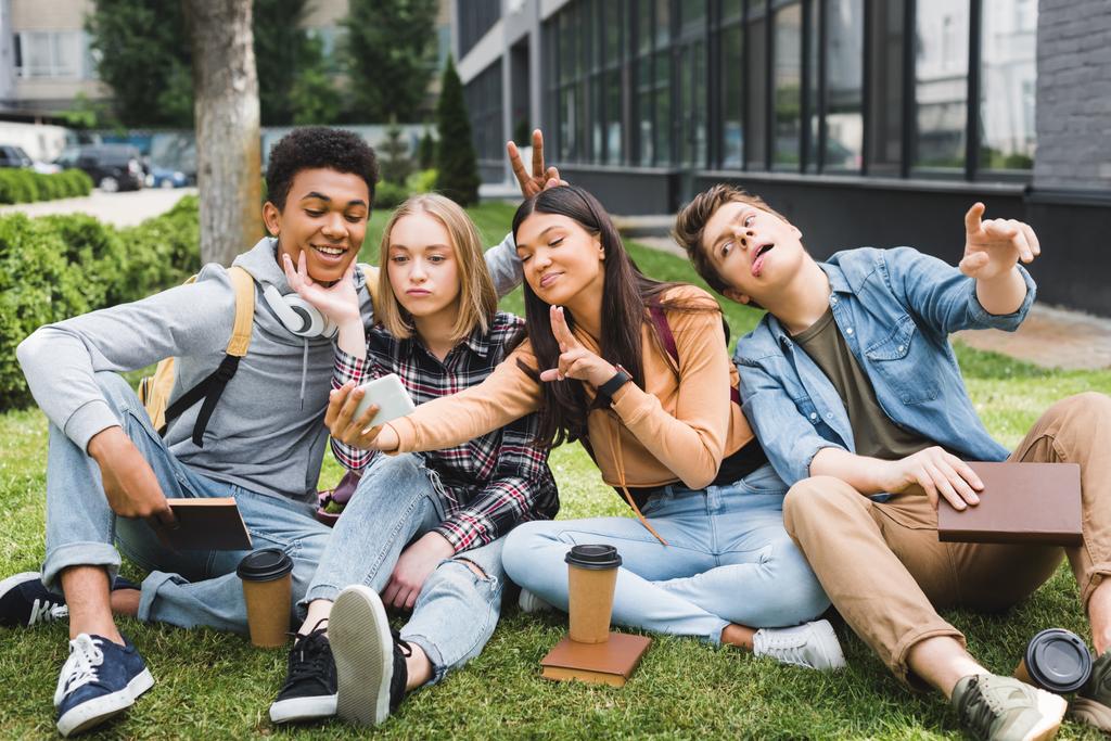 smiling and happy teenagers sitting on grass and taking selfie  - Photo, Image