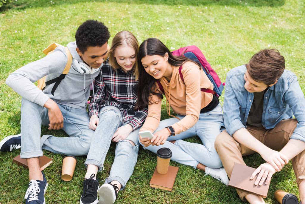 adolescents souriants et heureux assis sur l'herbe et regardant smartphone
  - Photo, image