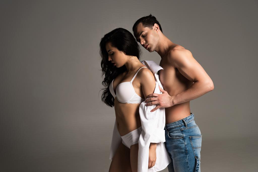 hemdloser muskulöser Mann in Jeans zieht sexy Freundin isoliert auf grau aus - Foto, Bild