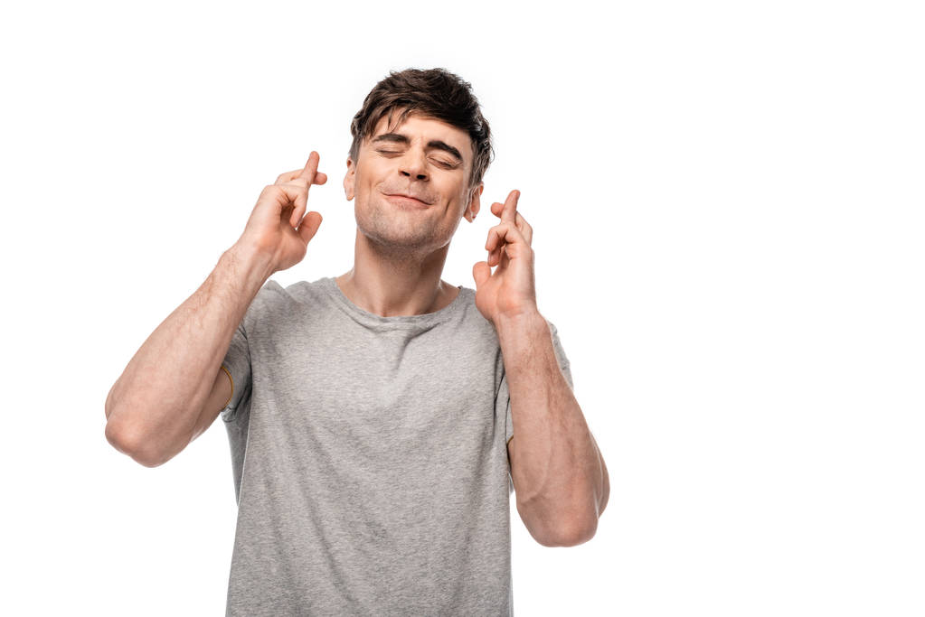 positieve jonge man Holding gekruiste vingers met gesloten ogen geïsoleerd op wit - Foto, afbeelding