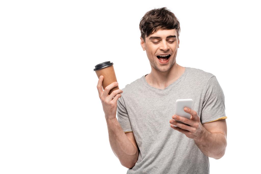 vrolijke jonge man met koffie te gaan tijdens het gebruik van smartphone geïsoleerd op wit - Foto, afbeelding