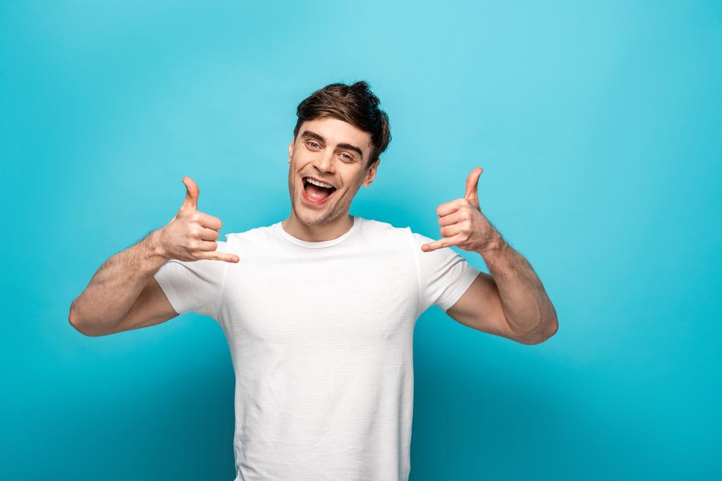 cheerful young man showing lets drink gestures while looking at camera on blue background - Photo, Image