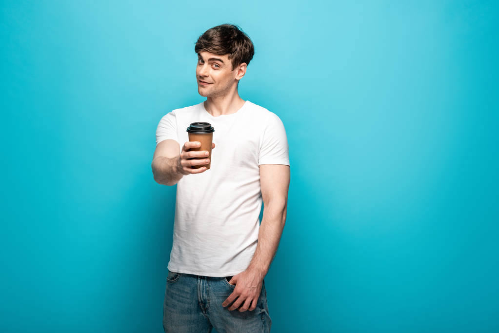 χαμογελαστός νέος άνθρωπος κρατώντας χάρτινο φλιτζάνι σε απλωμένο χέρι και κοιτάζοντας την κάμερα σε μπλε φόντο - Φωτογραφία, εικόνα