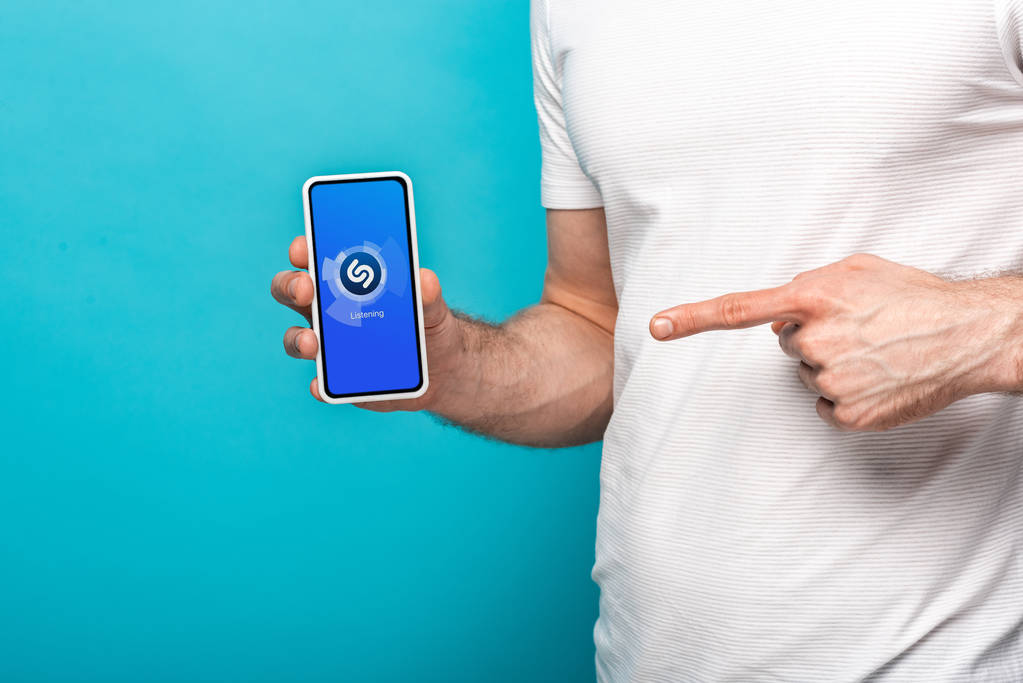 キエフ、ウクライナ - 2019年5月16日:Shazamアプリでスマートフォンを指す男のトリミングビュー、青で隔離 - 写真・画像