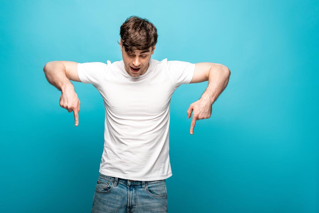 mladý muž v bílém tričku směřující dolů s prsty na modrém pozadí - Fotografie, Obrázek