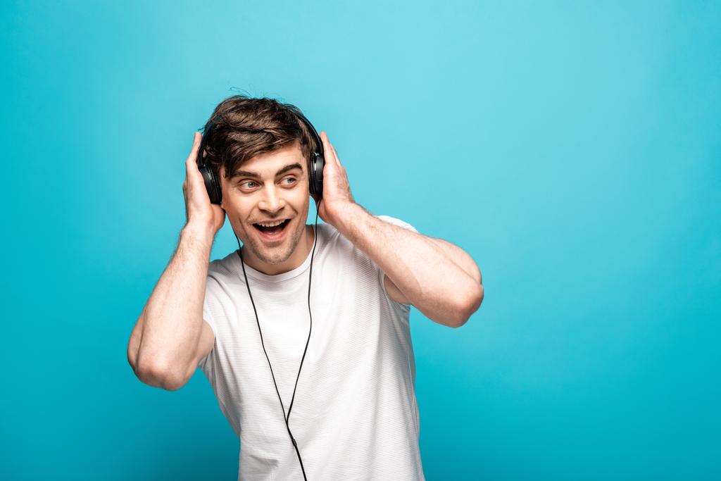 glimlachend jong man luisteren muziek in koptelefoon en op zoek weg op blauwe achtergrond - Foto, afbeelding