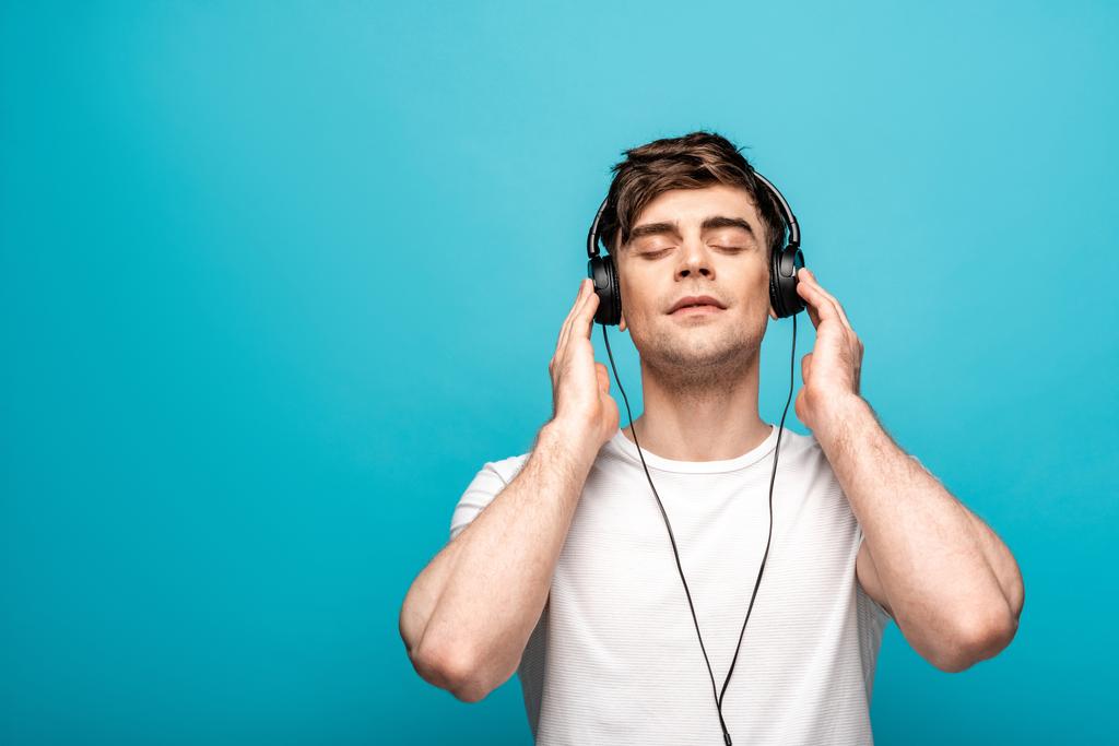 verträumter junger Mann mit Kopfhörern, der mit geschlossenen Augen auf blauem Hintergrund Musik hört - Foto, Bild