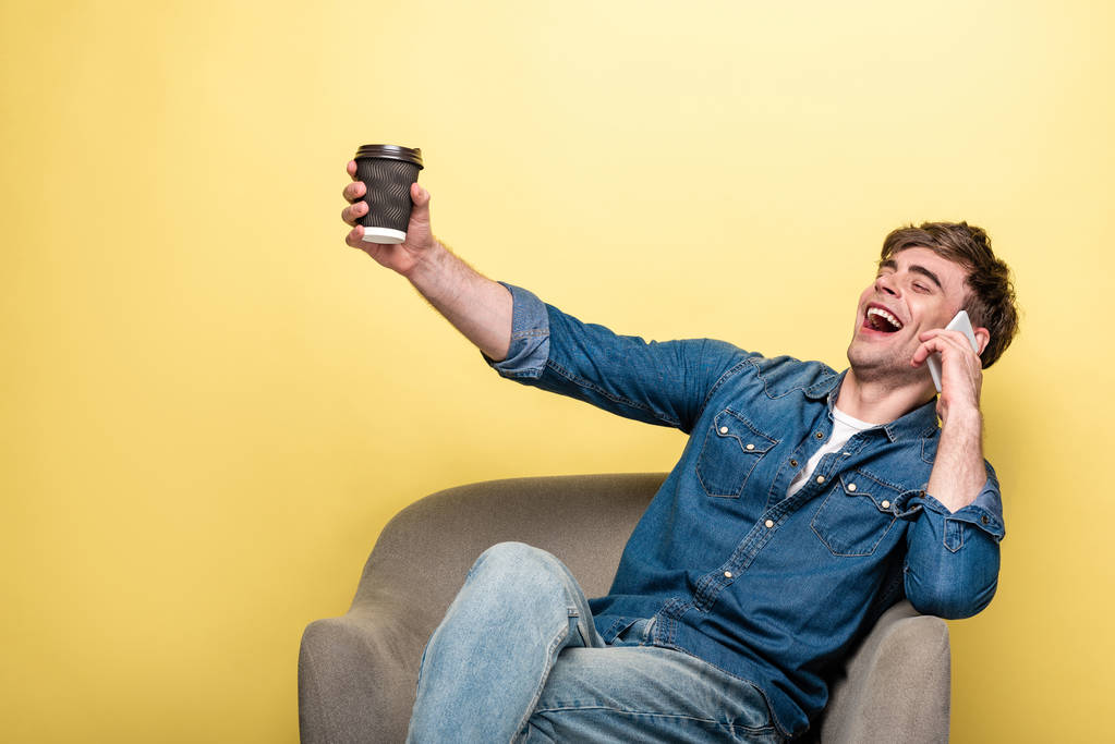 excited young man hoding paper cup while sitting in armchair and talking on smartphone on yellow background - Photo, Image
