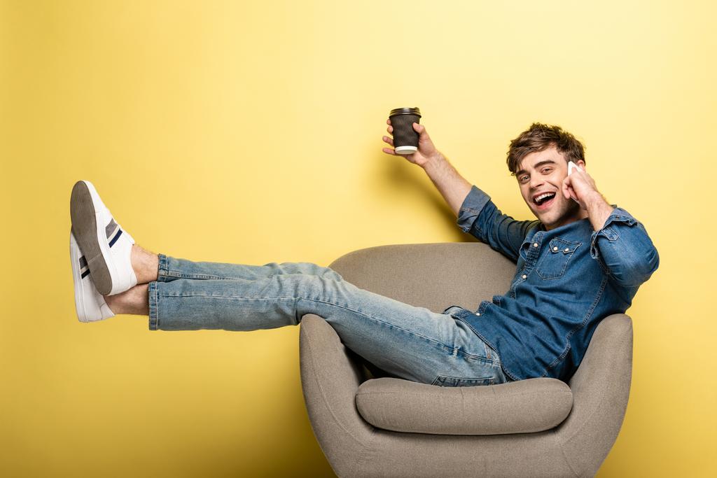 homem feliz sentado em poltrona enquanto fala no smartphone e segurando copo de papel no fundo amarelo
 - Foto, Imagem