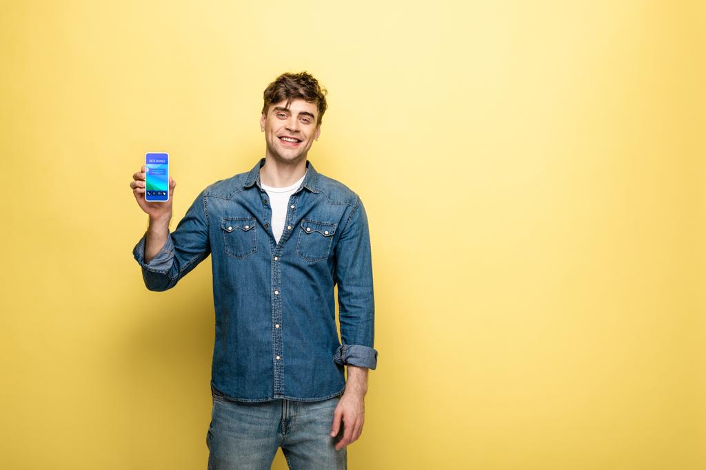 schöner lächelnder Mann in Jeanskleidung hält Smartphone mit Buchungs-App auf gelb - Foto, Bild