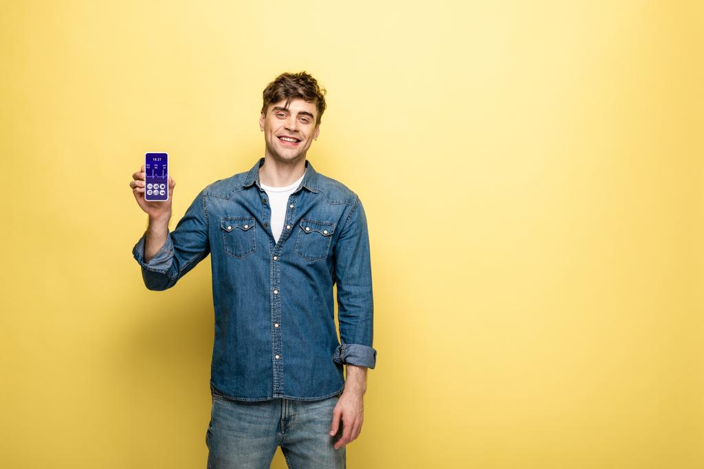 красивый улыбающийся мужчина в джинсовой одежде держит смартфон с приложением для здоровья на желтом
 - Фото, изображение