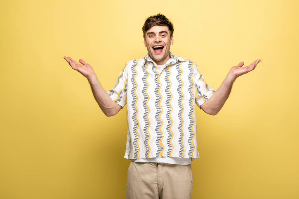 vrolijke jonge man toont schouderophalend gebaar en glimlachend op de camera op gele achtergrond - Foto, afbeelding