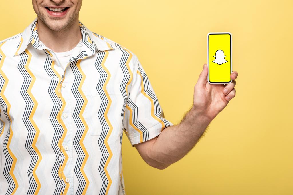 キエフ、ウクライナ - 2019年5月16日:黄色に隔離されたSnapchatアプリでスマートフォンを示す笑顔の男のトリミングビュー - 写真・画像