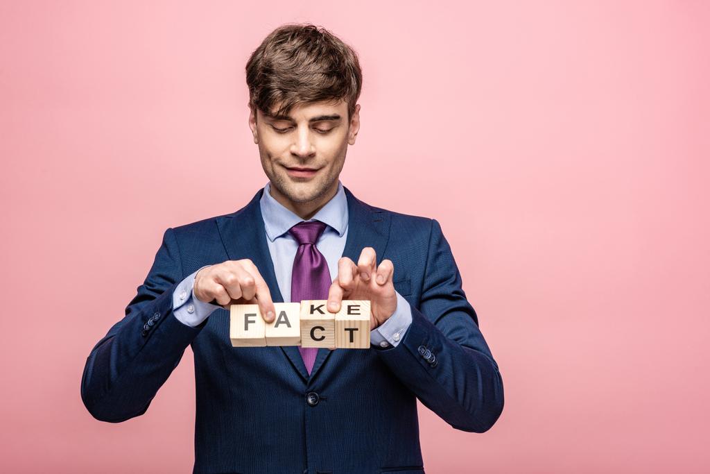 smiling man holding wooden cubes with fake fact inscription isolated on pink - Photo, Image