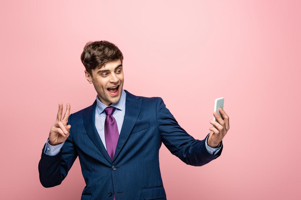 χαρούμενος επιχειρηματίας δείχνοντας χειρονομία νίκης, ενώ χρησιμοποιώντας το smartphone σε ροζ φόντο - Φωτογραφία, εικόνα