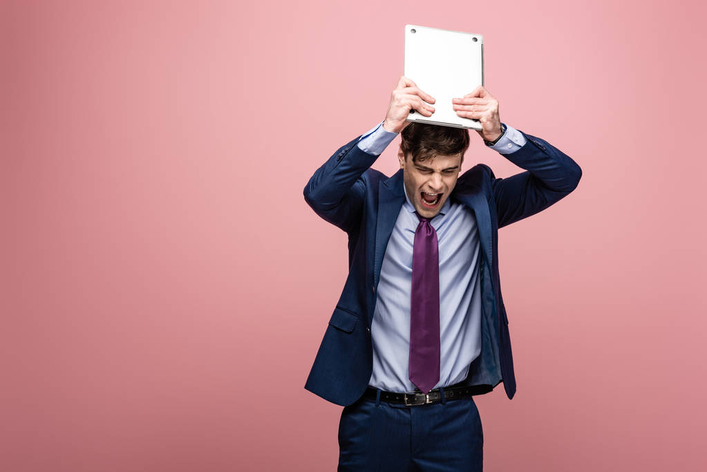 angry businessman quarreling while holding laptop above head on pink background - Photo, Image
