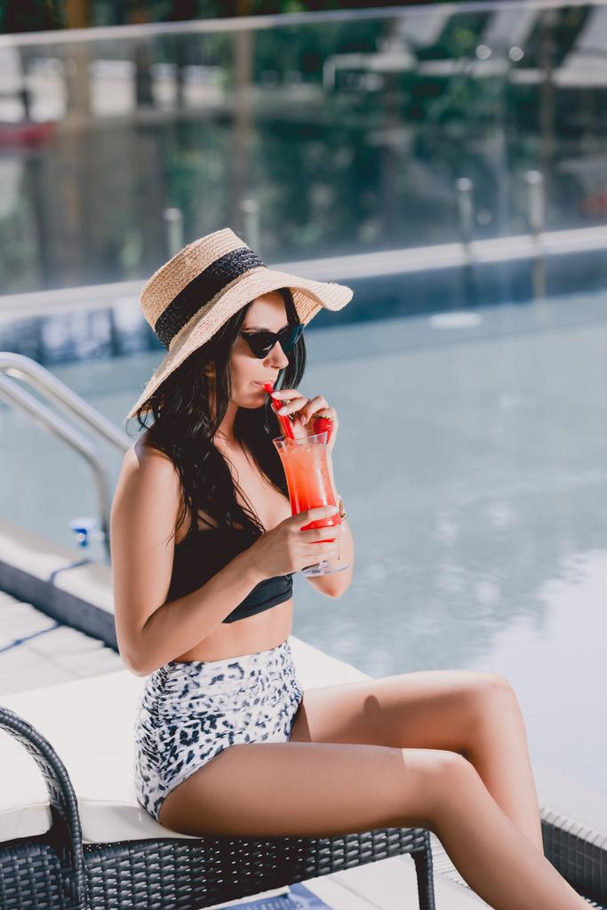 όμορφη μελαχρινή νεαρή γυναίκα σε μαγιό, ψάθινο καπέλο και γυαλιά ηλίου πίνοντας κοκτέιλ ενώ κάθονται σε ξαπλώστρες κοντά στην πισίνα - Φωτογραφία, εικόνα