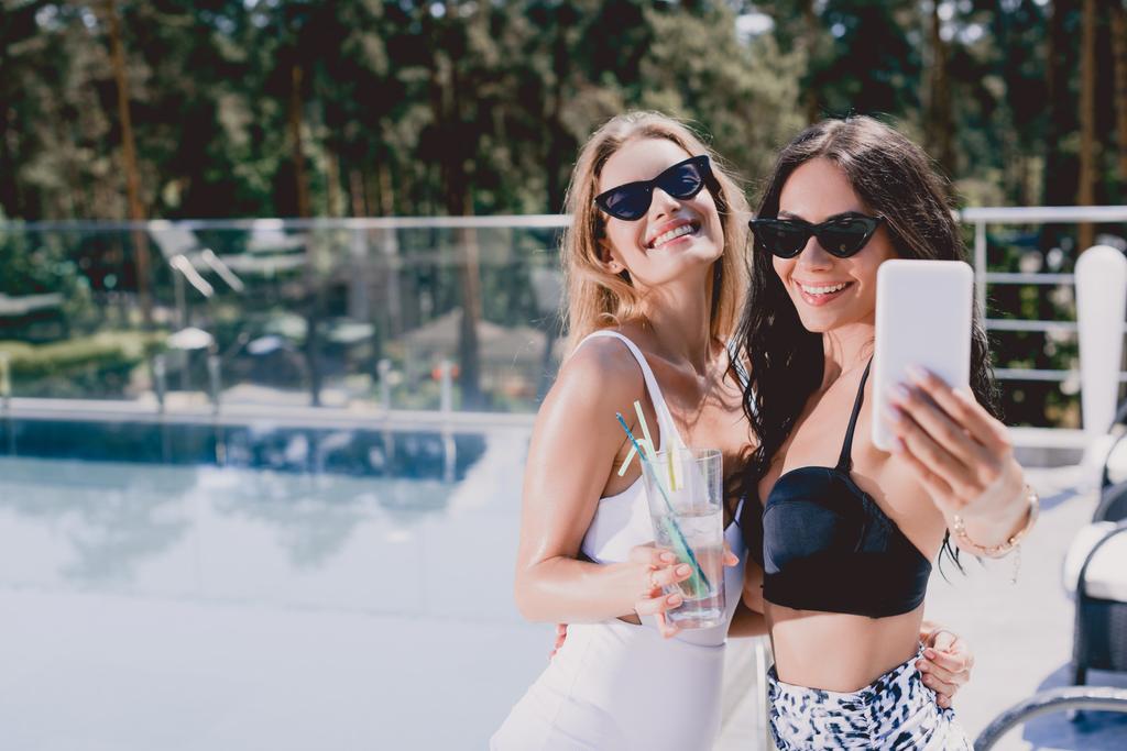 sexy et souriante brune et blonde femmes en maillots de bain prendre selfie près de la piscine
 - Photo, image